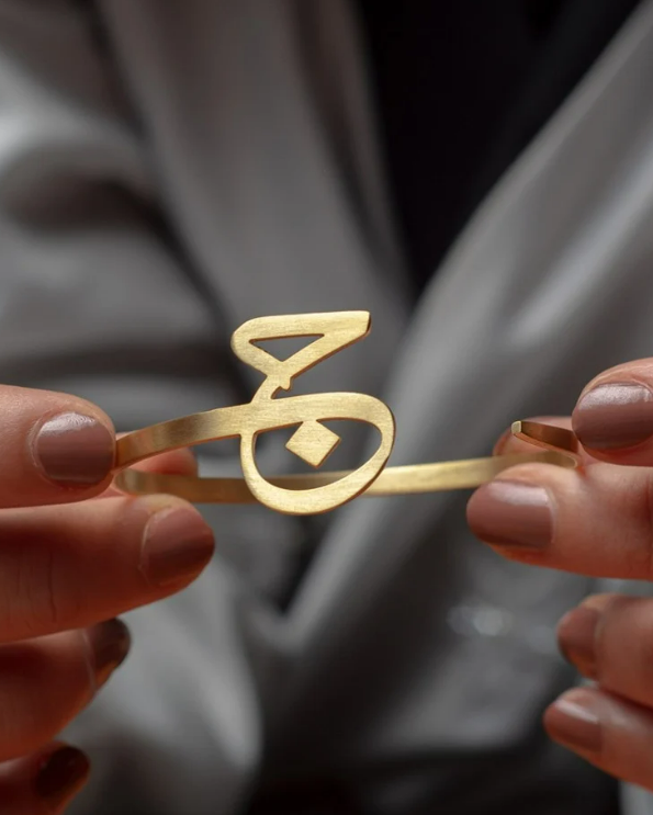 Gold hair clip with Arabic calligraphy held between two hands against a neutral background