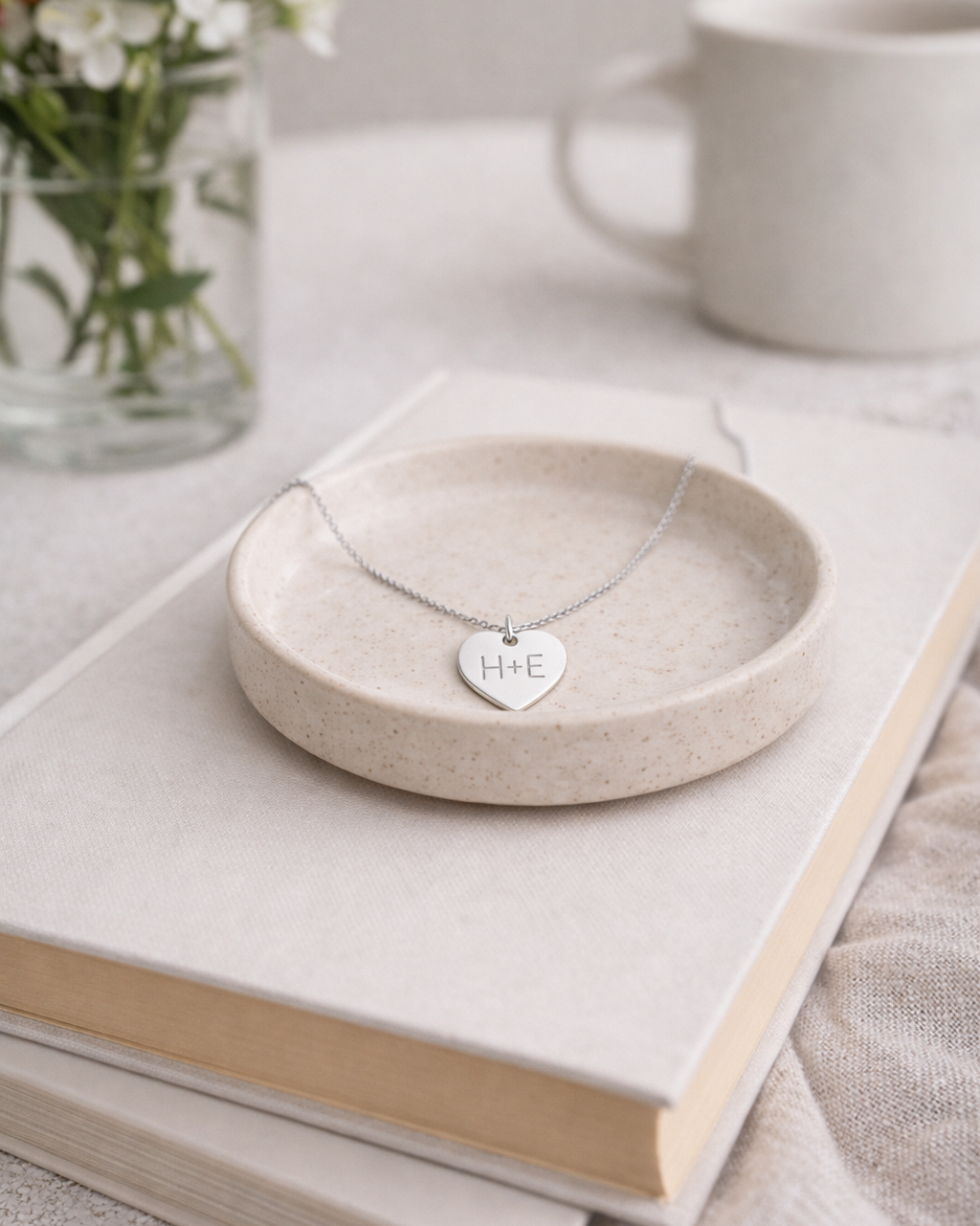 Necklace with heart-shaped pendant on a beige dish over books, with flowers and a teapot in the background.