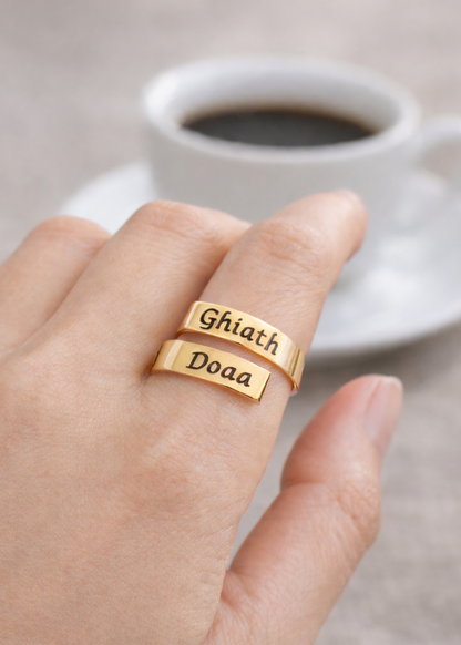 Gold ring with engraved names on a hand, with a cup of coffee and flowers in the background.