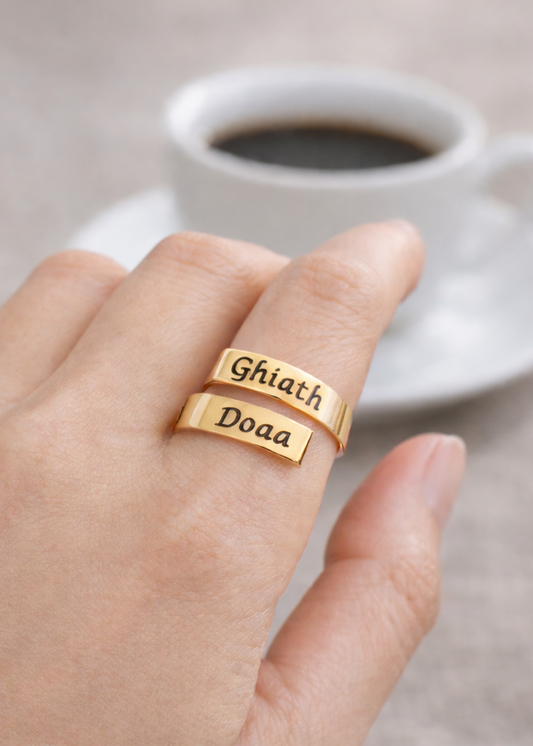 Gold ring with engraved names on a hand, with a cup of coffee and flowers in the background.