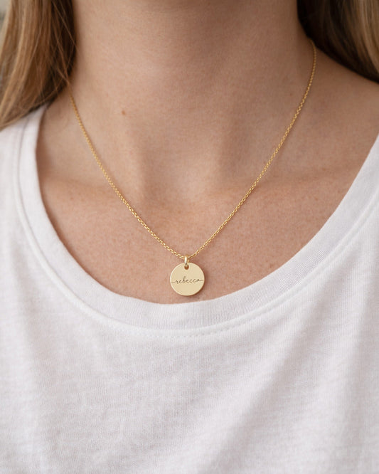 Person wearing a gold necklace with a round pendant on a white shirt.